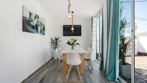 Een eetgedeelte met een witte tafel, stoelen, een hanglamp, een flatscreen televisie en potplanten.