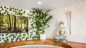 Een hottub met houten rand, een Boeddhabeeld en een muur met groene begroeiing en een boslandschap.