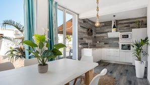 Een moderne keuken met een wit aanrecht, een ingebouwde oven en een magnetron. Er is een eethoek met een witte tafel en stoelen, en een potplant op de tafel.