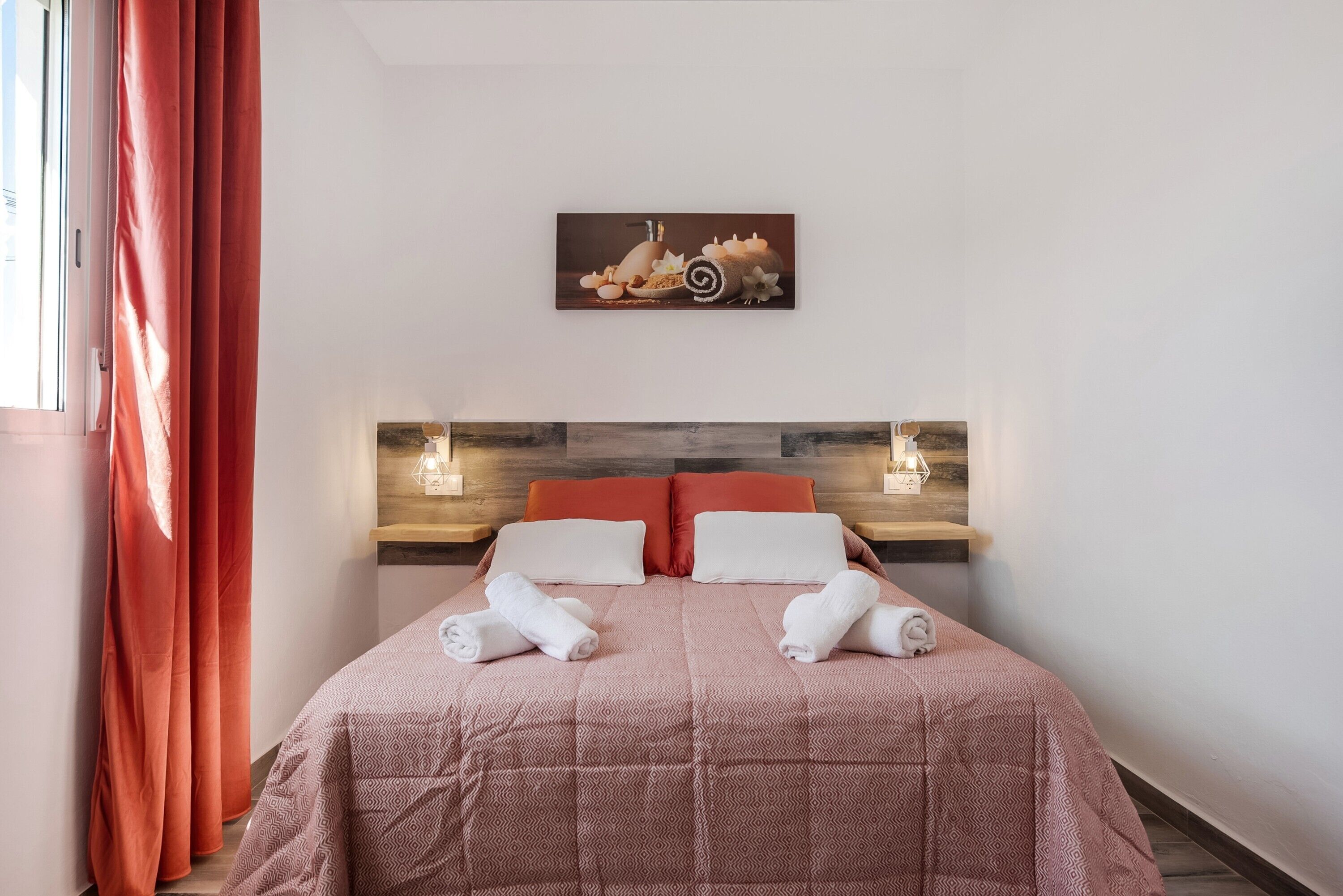Una cama bien hecha con almohadas, cabecera con luces fijadas a la pared y un cuadro encima.