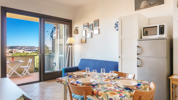 A dining area with a table set for four, a sofa, a refrigerator, and a view through a glass door.