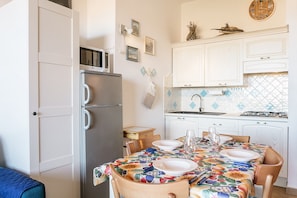 Une cuisine avec des meubles blancs, un four à micro-ondes, un réfrigérateur, une table à manger pour deux personnes et une horloge au mur.