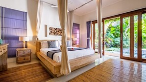 A spacious bedroom with a large bed, wooden headboard, and a canopy. There is a nightstand with a lamp and a wooden dresser. The room has large windows with curtains and a view of greenery outside.