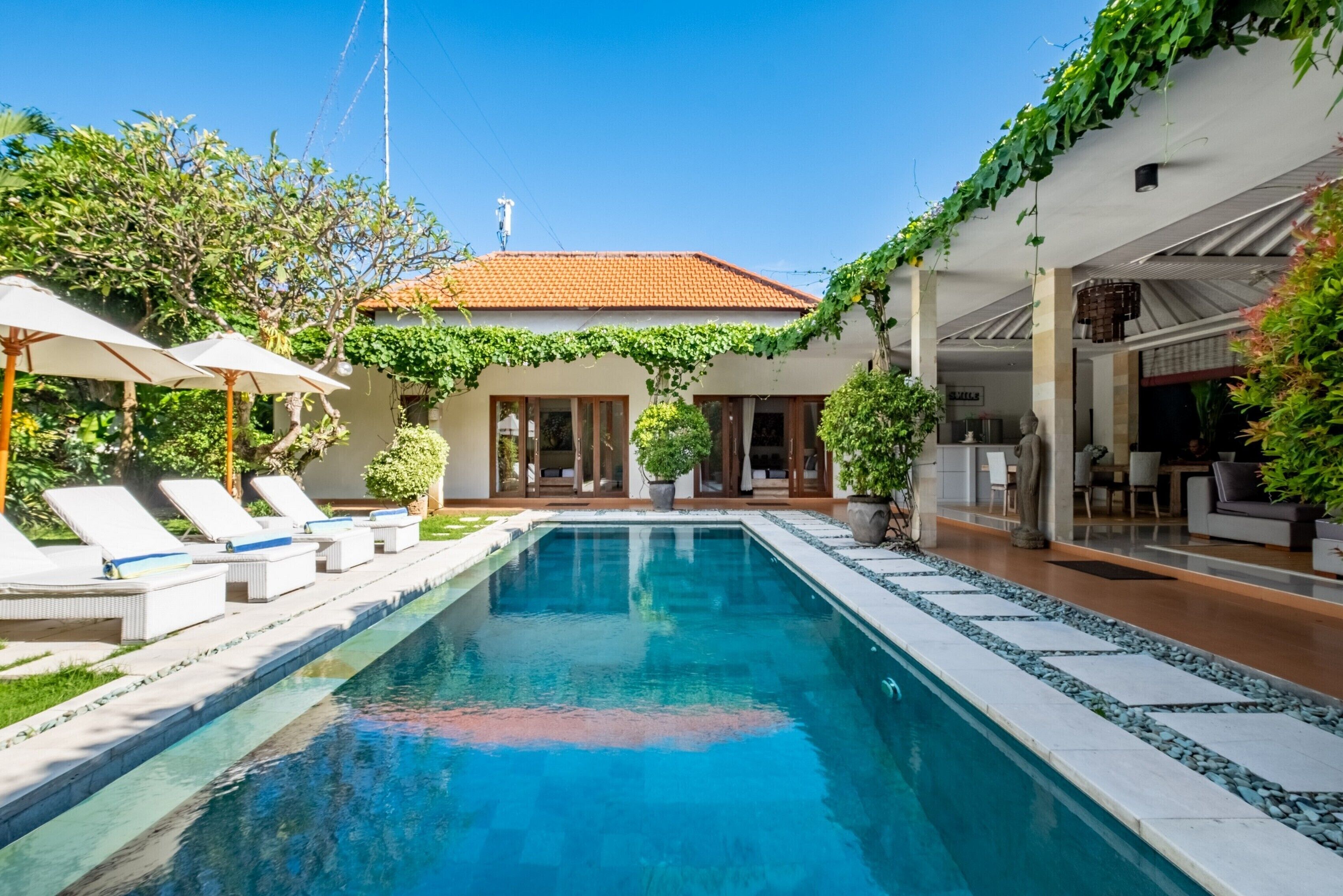 A swimming pool with lounge chairs, a covered patio, and a building with a tiled roof.