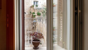 A balcony with a potted plant, a metal railing, and a view of a stone building with windows and greenery. - Vico Delle Palme (Monopoli)