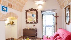 A cozy living room with a red sofa, a wooden coffee table with fruits, a small table with decorative items, a mirror, and a window with a net curtain. - Vico Delle Palme (Monopoli)