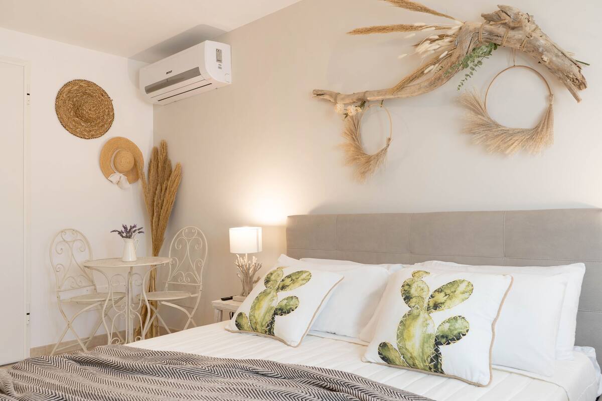 Une chambre à coucher comprenant un lit, deux chaises, une petite table et des éléments de décoration murale, notamment un coussin en forme de cactus et une tapisserie murale tissée.