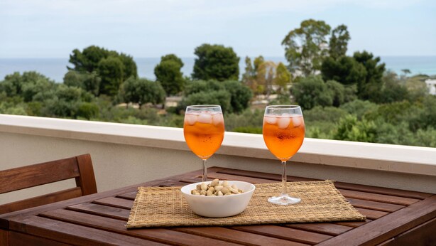 A wooden table with two glasses of orange beverage and a bowl of nuts on a balcony overlooking a scenic view of trees and the sea. - Suite Martino Seaview (Monopoli)