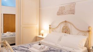 A neatly made bed with a blue patterned bedspread, white pillows, and a small floral decoration. There is a bedside table with a lamp and a wall-mounted picture. - Donna Lisa (Polignano a Mare)