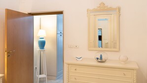 A room with a wooden door, a white dresser with drawers, a mirror, and a blue lamp on a white stool. - Donna Lisa (Polignano a Mare)