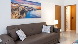 A modern living room with a brown sofa, patterned pillows, a side table with a lamp, and a large coastal landscape painting on the wall. - Donna Lisa (Polignano a Mare)