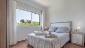 A bedroom with a bed, a nightstand, a window with curtains, and a view of greenery outside. - Villa with pool and sea view - Villa Rica (MENORCA)