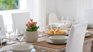 A dining table set with white plates, bowls, and wine glasses, a potted plant with pink flowers, and a banana on a wooden board. - Villa with pool and sea view - Villa Rica (MENORCA)