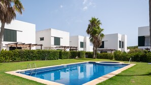 A swimming pool with a wooden deck, surrounded by manicured grass and hedges, with modern white buildings in the background. - Villa with pool and sea view - Villa Rica (MENORCA)