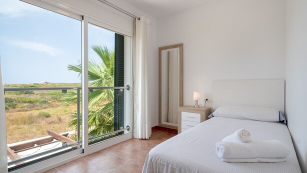 A bedroom with a large sliding glass door, a bed with a white bedspread, a nightstand with a lamp, and a view of the landscape. - Villa with pool and sea view - Villa Rica (MENORCA)