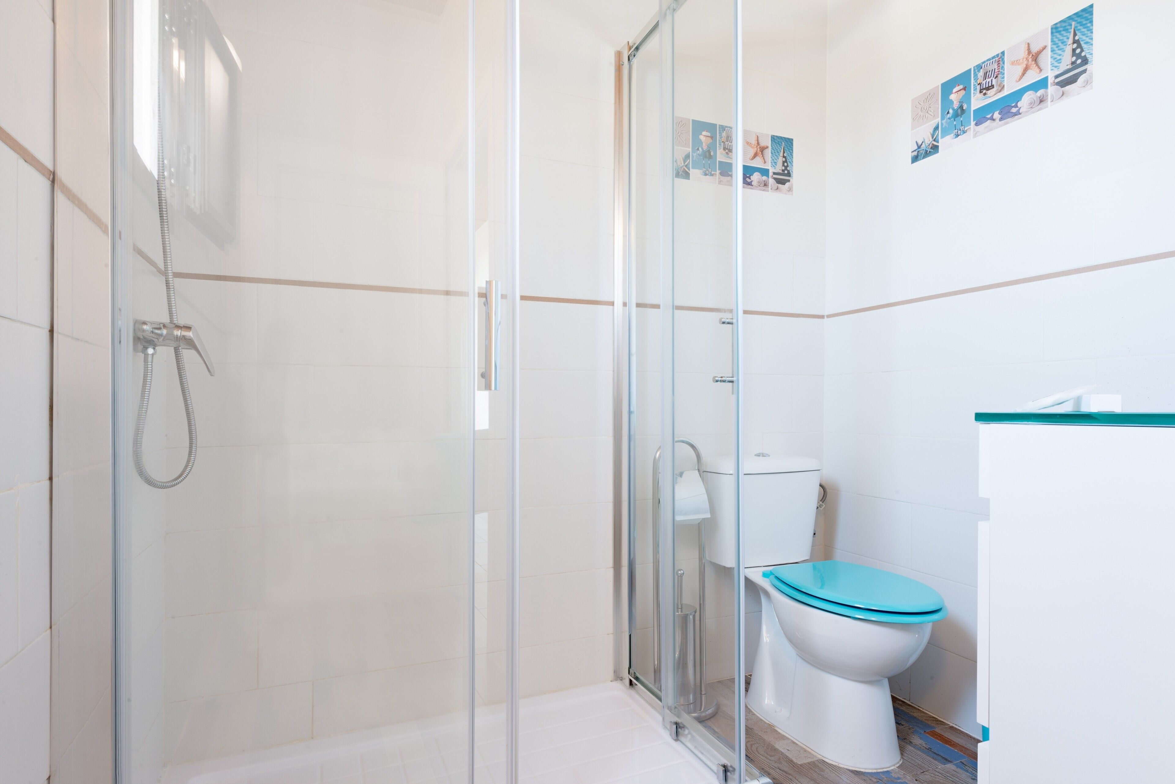 Een badkamer met een douchecabine van glas, een toilet met een blauwe zitting, en een muur met decoraties in nautisch thema.