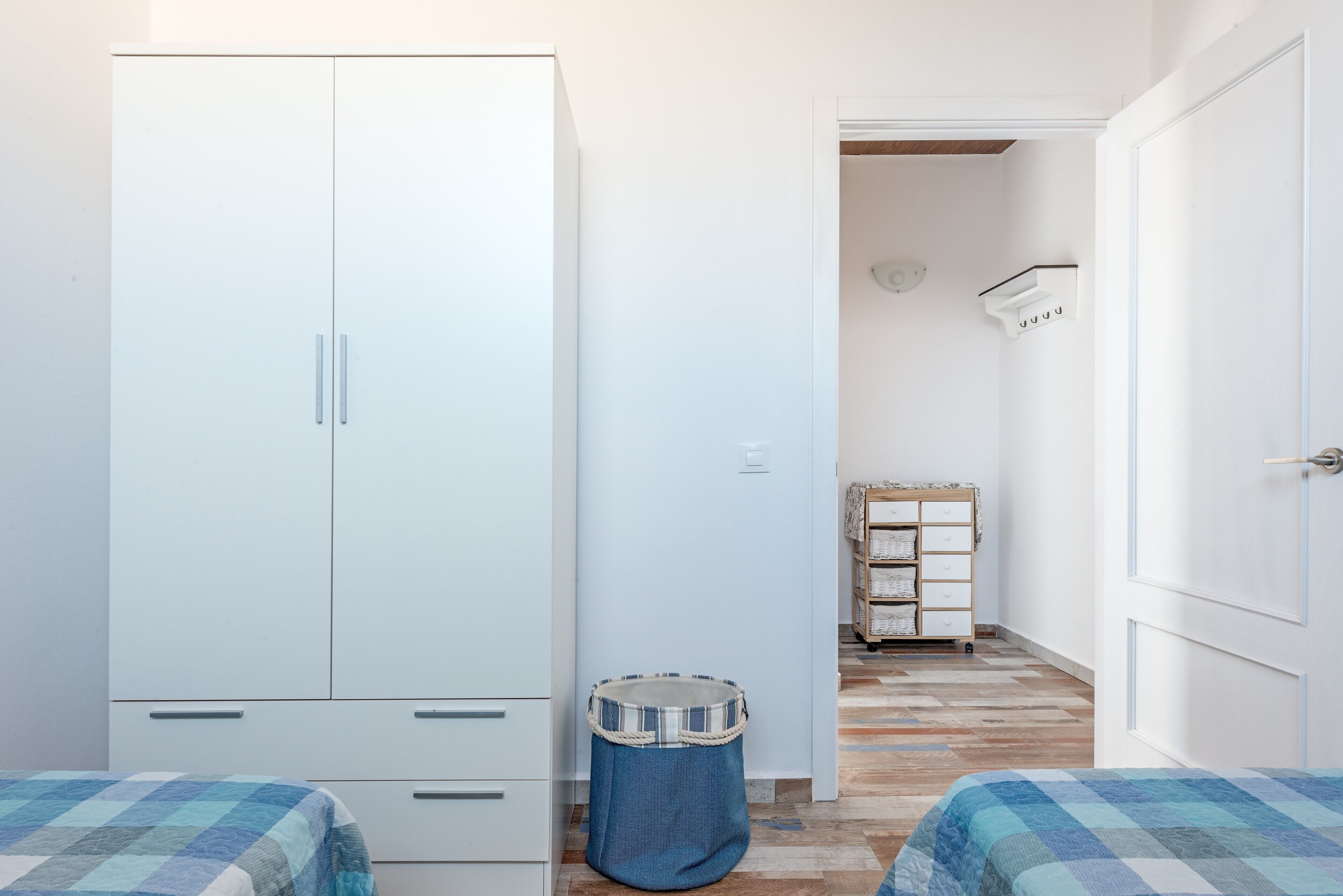A bedroom with a wardrobe, a pet bed, a small wooden cabinet, and a bed with a checkered bedspread.