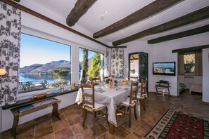 A dining area with a table set for four, large windows with a view of a lake and mountains, and a television mounted on the wall.