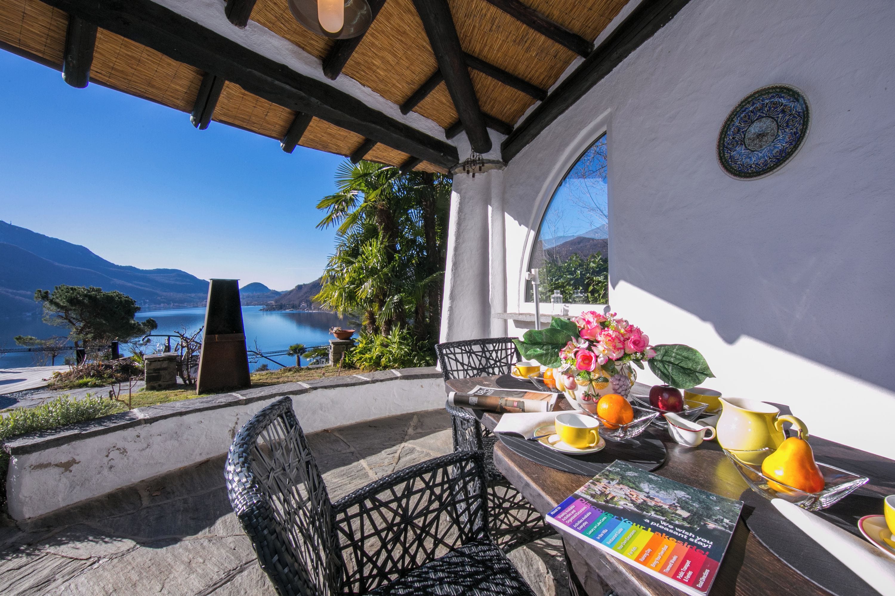 A terrace with a view of a lake and mountains, a table with breakfast items, and a chair.