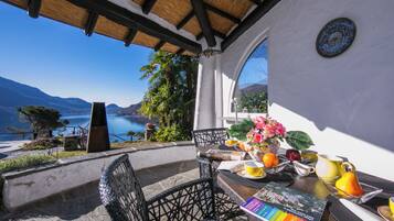 A terrace with a view of a lake and mountains, a table with breakfast items, and a chair.