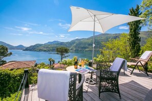 A deck with outdoor furniture, a table with a bottle and glasses, and a view of a lake and mountains. - Morcote Magic View (Morcote)