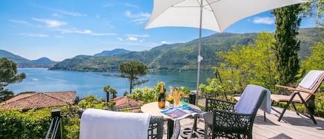 Una terraza con mobiliario exterior, una mesa con una botella y vasos, y vistas a un lago y montañas.