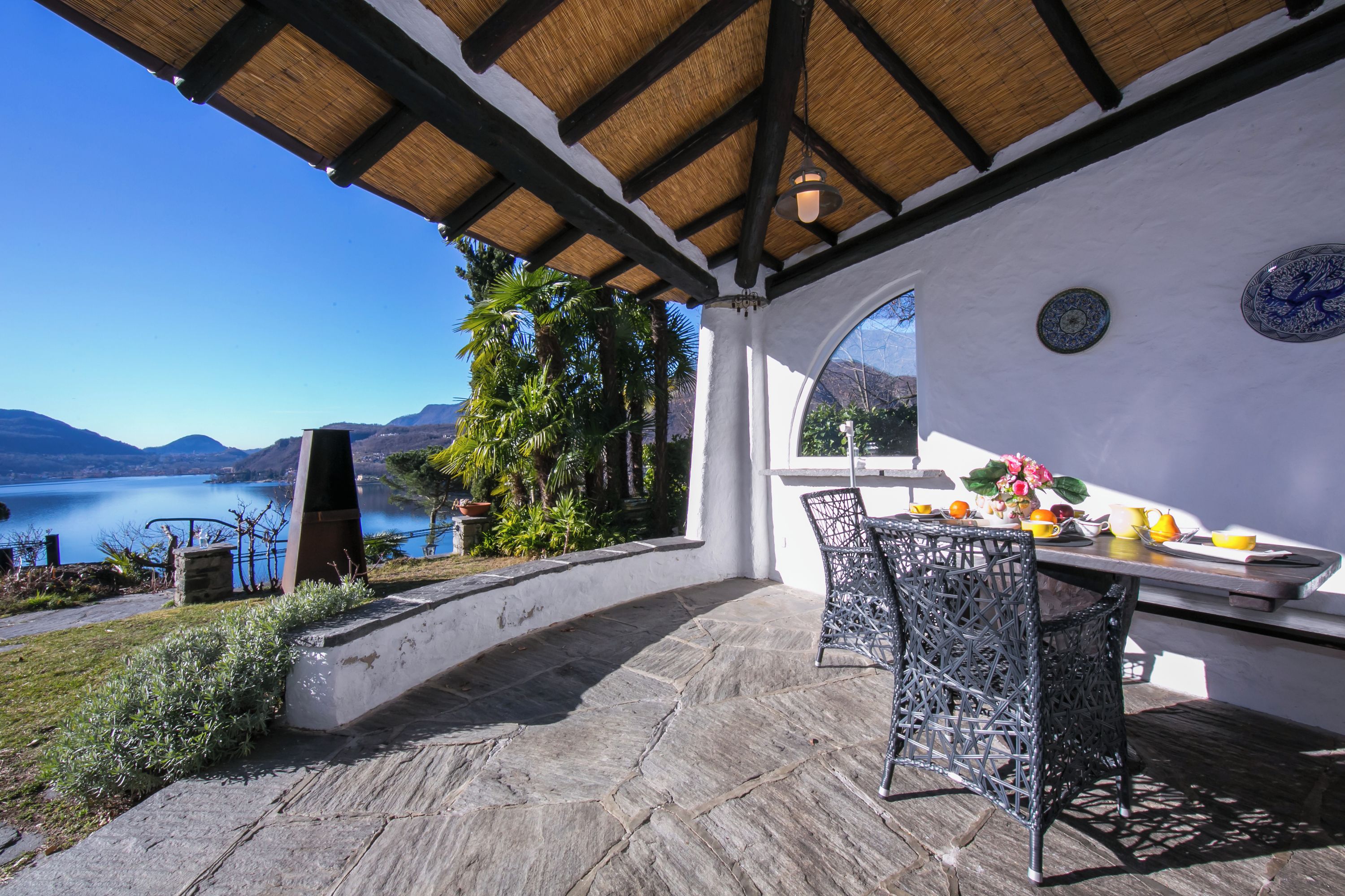 Un patio al aire libre con piso de piedra, una mesa con sillas, vista a un lago y una estructura de techo de paja.
