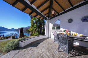 An outdoor patio with a stone floor, a table with chairs, a view of a lake, and a thatched roof structure. - Morcote Magic View (Morcote)