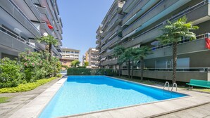 A swimming pool surrounded by apartment buildings, with a bench and palm trees nearby.