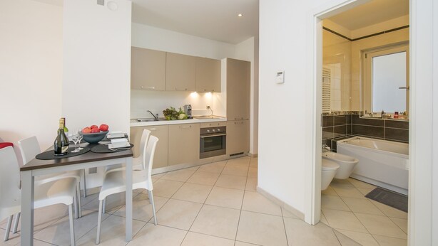 A modern kitchen with white cabinets, a dining table with chairs, and a small island with a bowl of fruit and a bottle of wine. - Prestige du Lac 4 (Lugano)