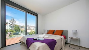 A bedroom with a large window, a bed with a purple blanket, and a side table with a lamp. - Urban Flat Paradiso (Paradiso)