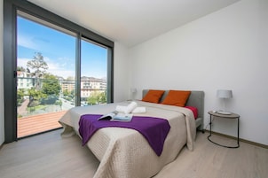 Quarto com uma janela grande, uma cama com cobertor roxo e uma mesa de cabeceira com abajur.