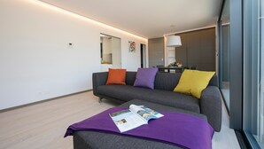A modern living room with a grey sofa, colorful cushions, a magazine on a purple ottoman, and a glass wall offering a view into the kitchen. - Urban Flat Paradiso (Paradiso)