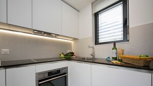 A modern kitchen with white cabinets, a built-in oven, a stainless steel range hood, a window with blinds, and a countertop with a bottle of wine, vegetables, and bread. - Urban Flat Paradiso (Paradiso)