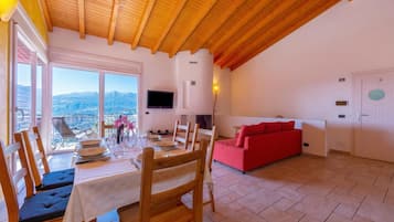 A dining area with a table set for four, wooden chairs, a red sofa, a TV mounted on the wall, and a view of mountains through large windows.
