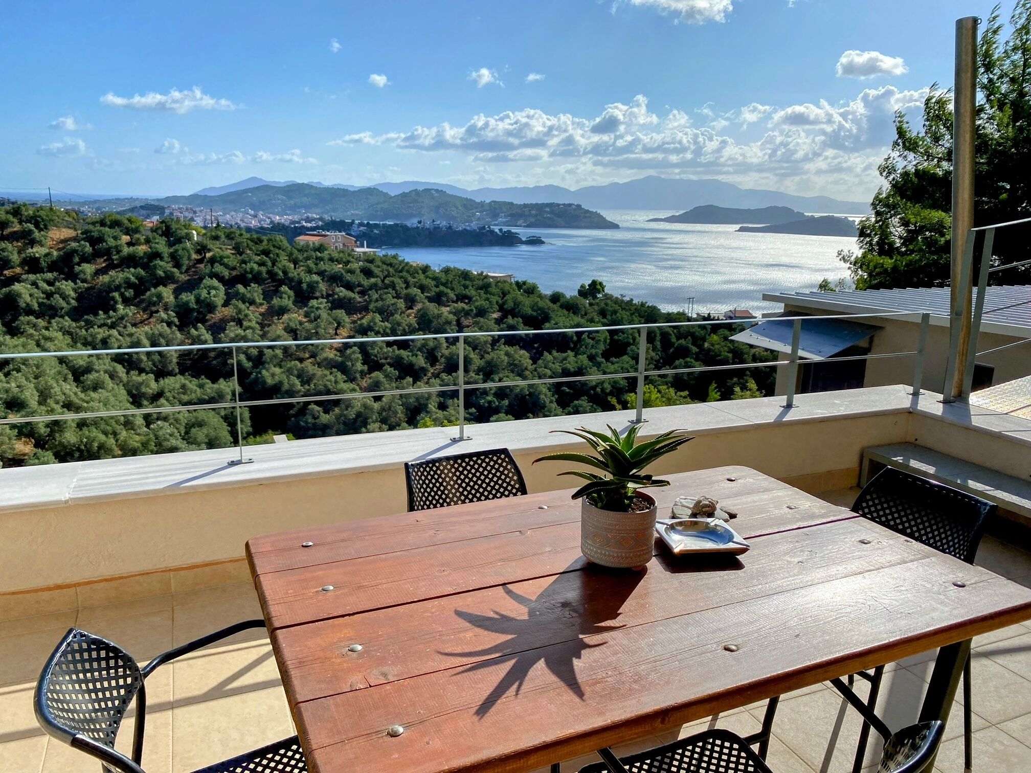 Een houten tafel met een potplant, een bord en een vork op een balkon met uitzicht op een kustlandschap met bergen en een waterlichaam.