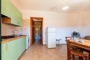 Une cuisine équipée d’une cuisinière, d’un évier, d’un lave-linge et d’un réfrigérateur. On y trouve une table à manger avec des chaises, ainsi qu’un téléviseur posé sur une petite table.