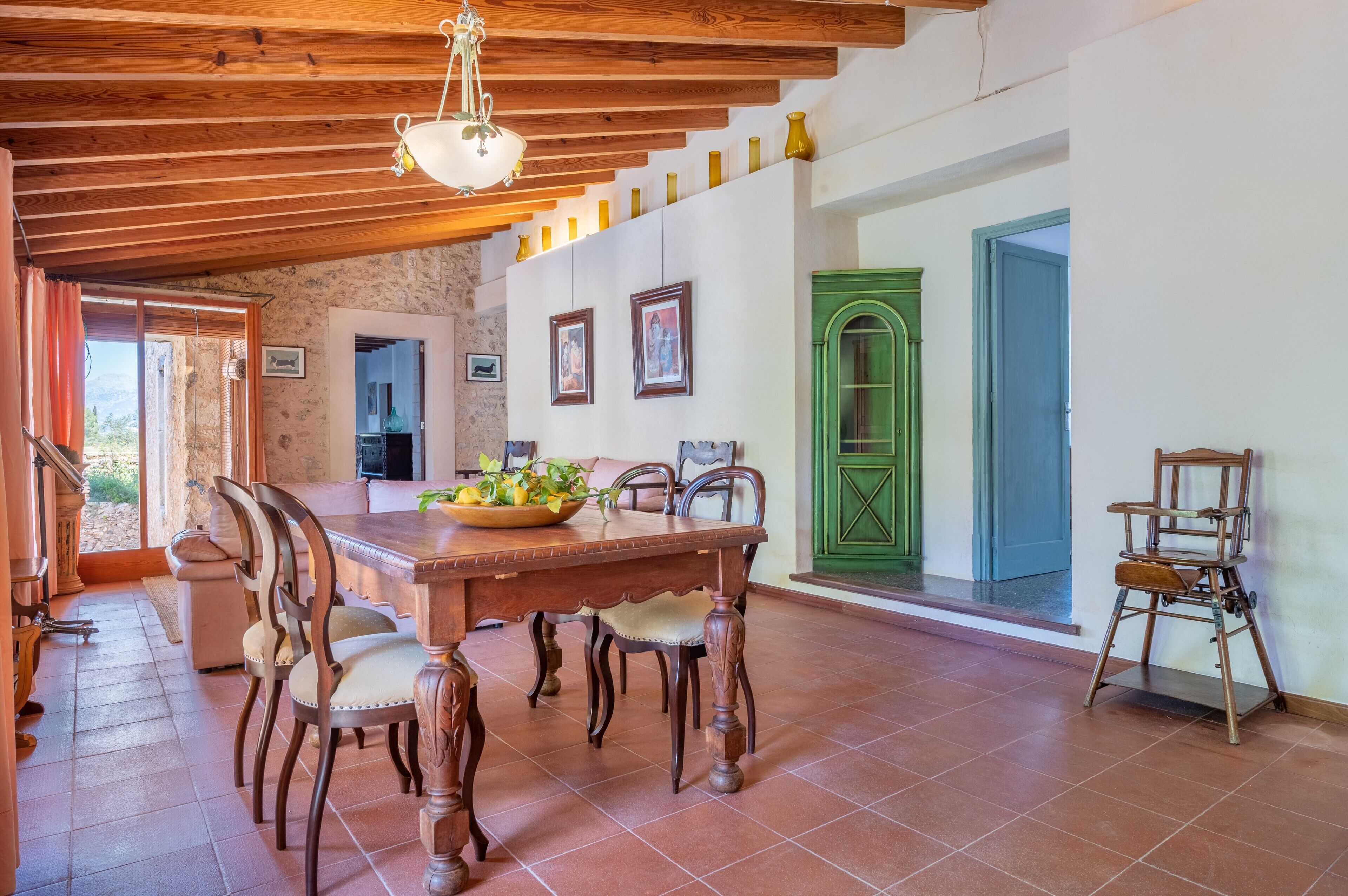 A dining area with a wooden table, chairs, and a bowl of fruit. There are framed pictures on the wall, a green door, and a staircase leading to another room.