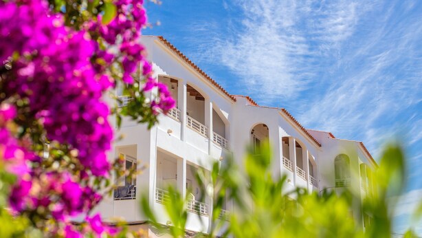 Un edificio blanco con varios balcones y ventanas arqueadas, rodeado de flores rosadas y vibrantes.