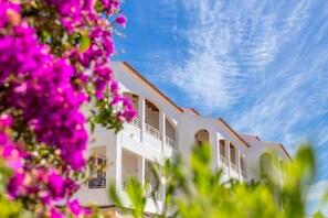 A white building with multiple balconies and arched windows, surrounded by vibrant pink flowers.
