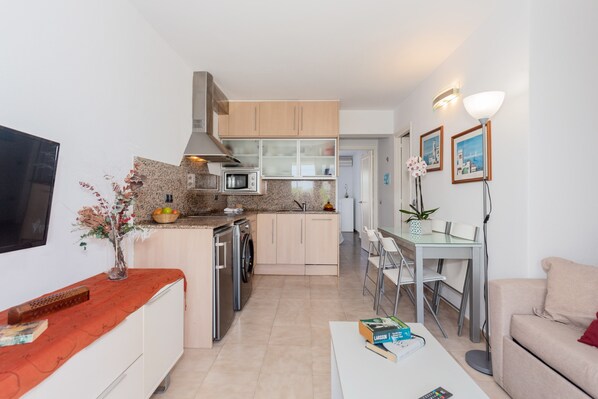 A modern kitchen with a microwave, oven, and dishwasher, a dining area with a table and chairs, and a living area with a sofa and a flat-screen TV.