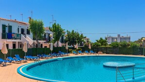 Una piscina con sillones reclinables azules, un deck con baldosas y un edificio con balcones al fondo.