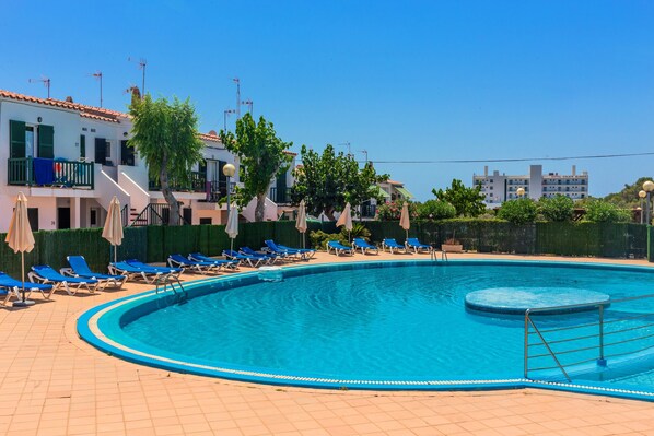 Una piscina con sillones reclinables azules, un deck con baldosas y un edificio con balcones al fondo.