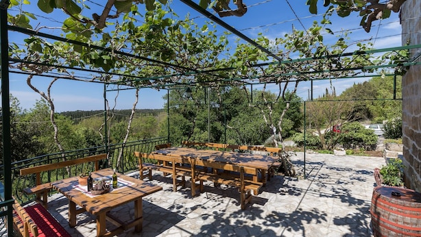 An outdoor patio with wooden tables and benches, surrounded by greenery and a stone floor. - Country House Bakicevo (Konavle)