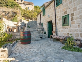 Un patio de piedra con un barril, un banco y un edificio con contraventanas verdes.