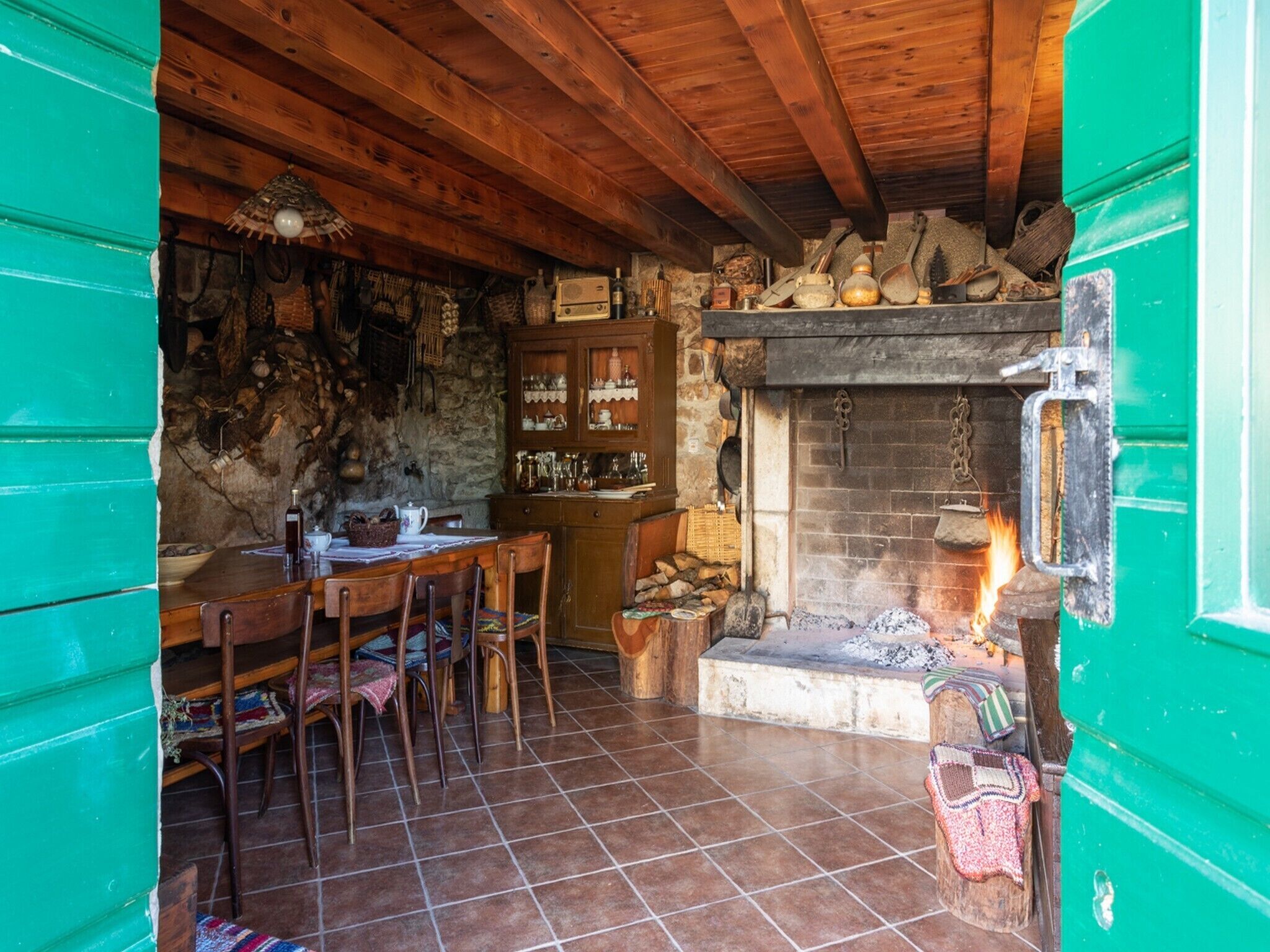 Un interior rústico con techo de madera, pared de piedra, chimenea y comedor con sillas y mesa.