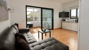 A living room with a sofa, a small table, and a kitchen area with a TV, microwave, and coffee maker. - Close to Hiking and Beach with Beautiful Winter Garden - Apartment Ye (Haria)