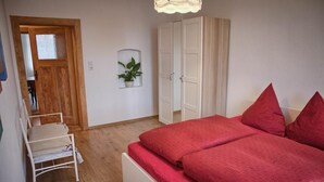 Une chambre avec une porte en bois, une armoire blanche, un lit avec une literie rouge, une chaise blanche et une plante en pot accrochée au mur.