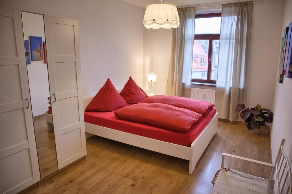 A bedroom with a white bed, red bedding, a bedside lamp, a window with curtains, and a potted plant.