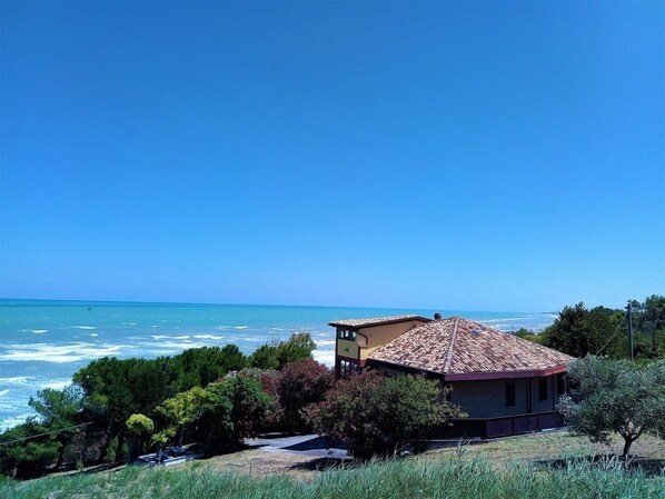 A coastal house with a tiled roof, surrounded by greenery and overlooking the sea.